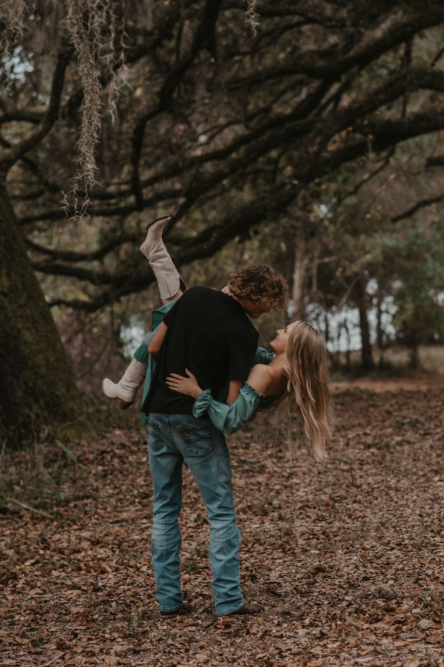 A couple embraces while standing among fallen autumn leaves in a wooded area during a romantic photoshoot.