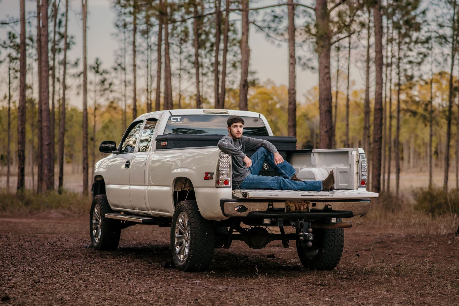 A lifted white pickup truck parked in a wooded area at sunset with someone relaxing in the truck bed.