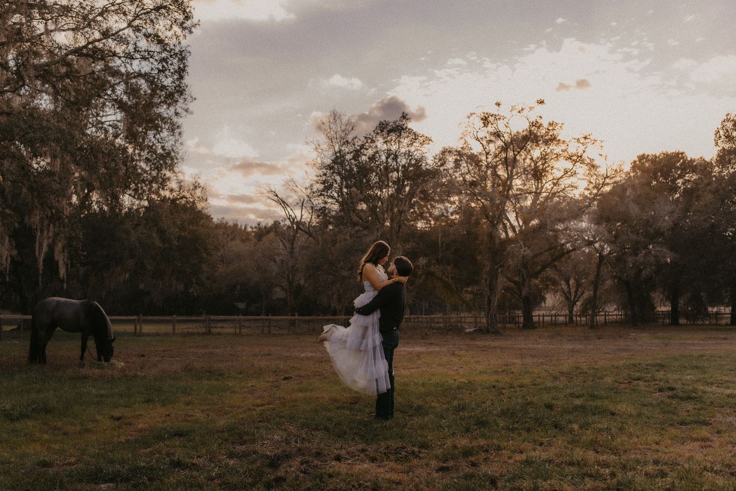 A couple dances in a rustic field at sunset with horses grazing in the background.