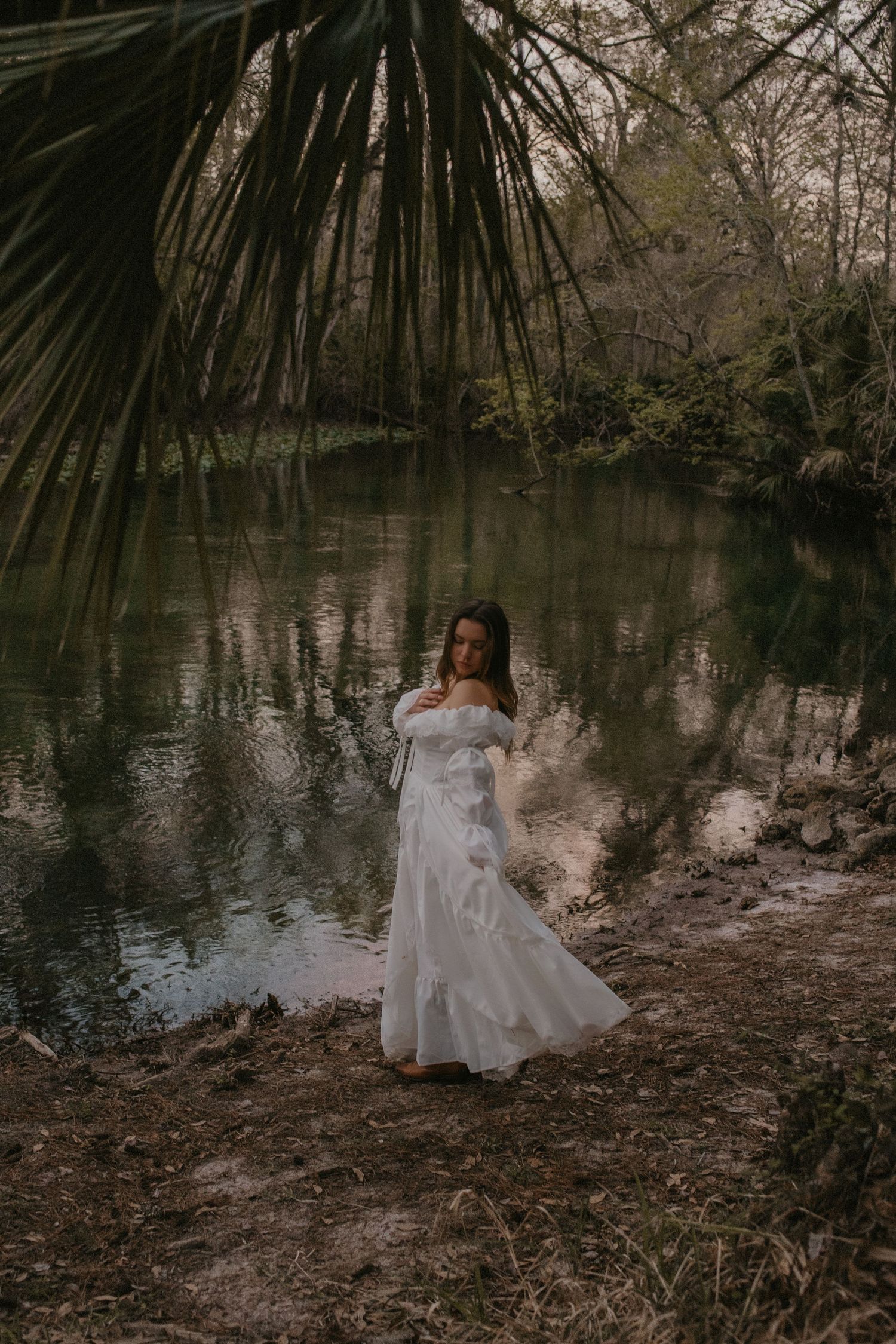 A figure in a flowing white dress sits contemplatively by a tranquil lake surrounded by palm fronds and trees at dusk.