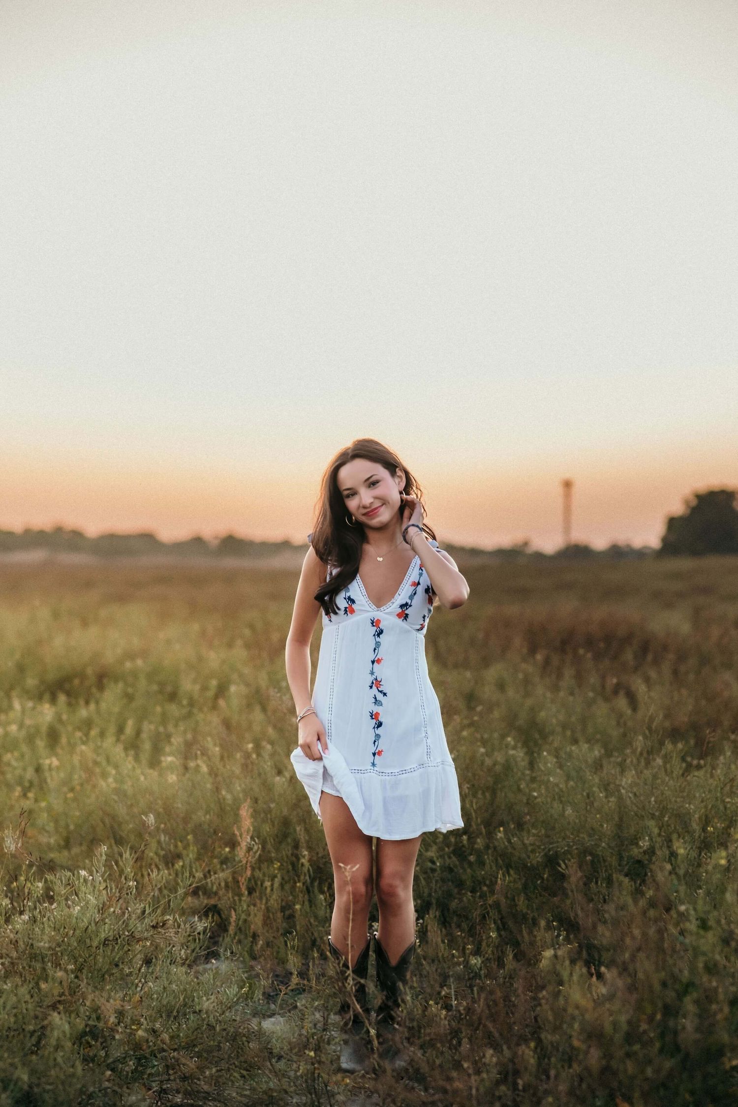 Someone in a white summer dress poses playfully in a rural field at sunset.