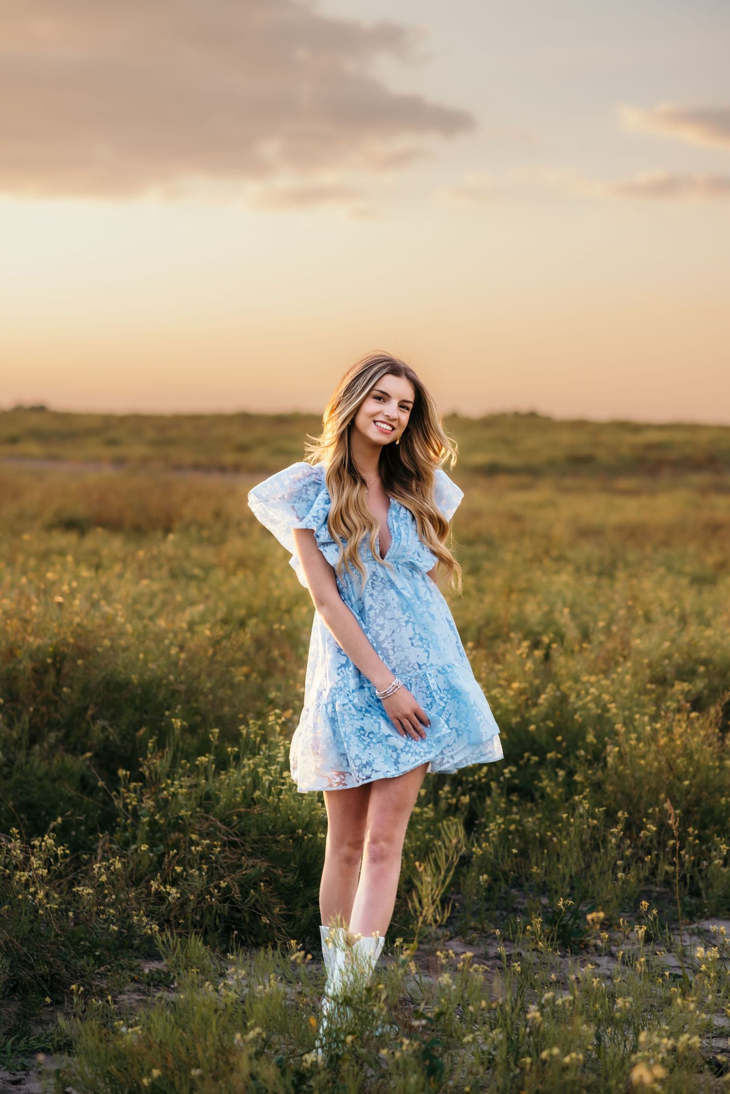 A woman in a blue dress and white boots stands among wildflowers during sunset.