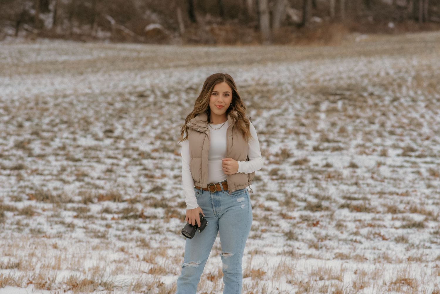 A person in light wash jeans and beige cardigan poses in a snowy winter field at sunset.