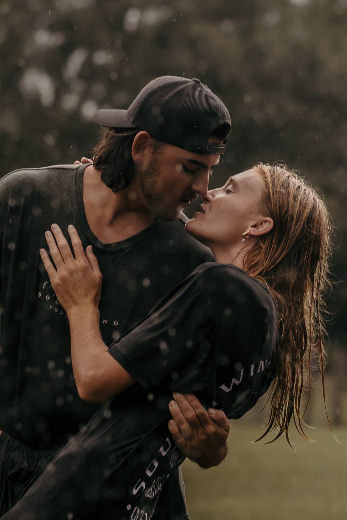 A couple shares an intimate moment in the rain while embracing and kissing in a moody outdoor setting.