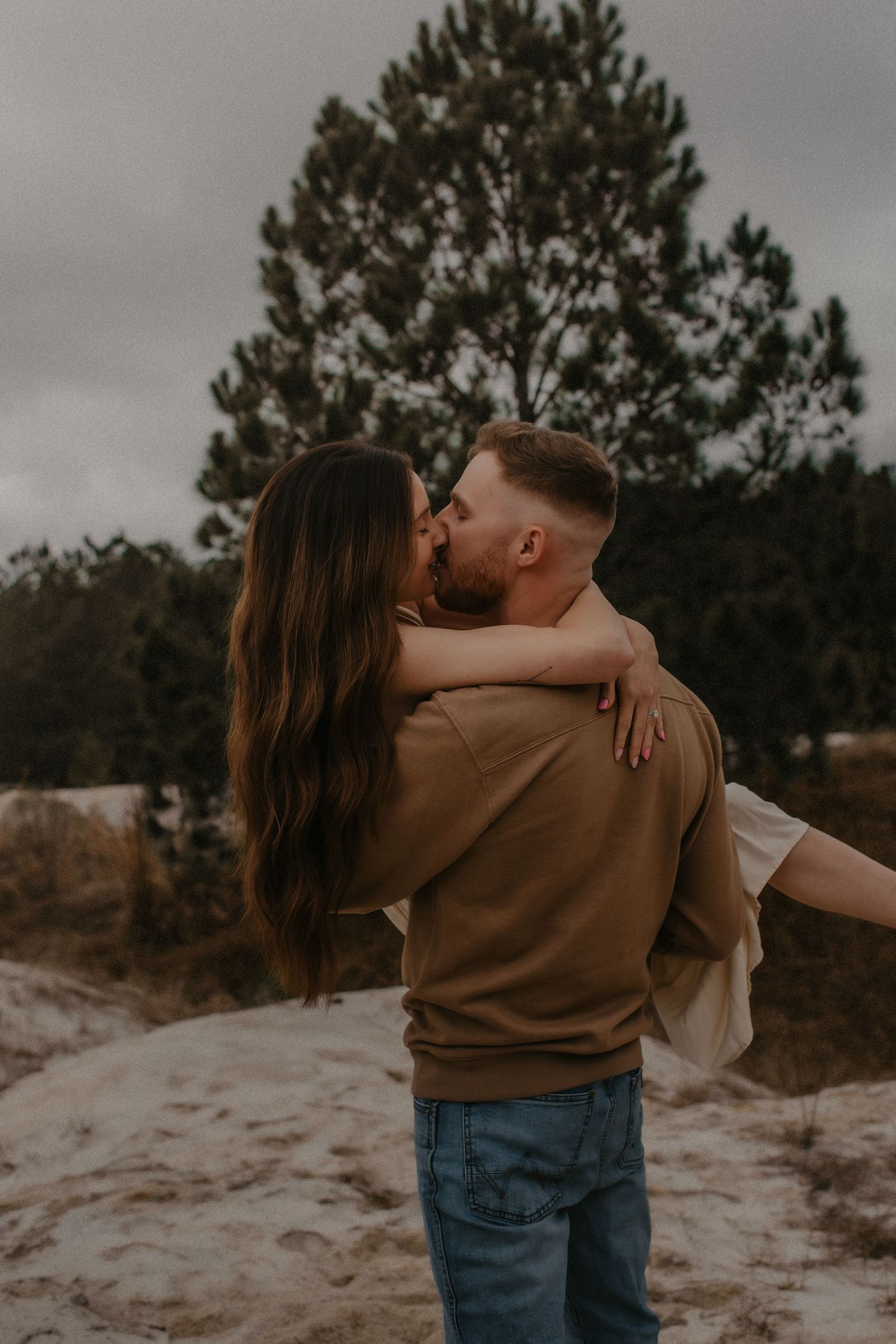 A couple embraces tenderly in a romantic outdoor photoshoot against a backdrop of pine trees and moody skies.