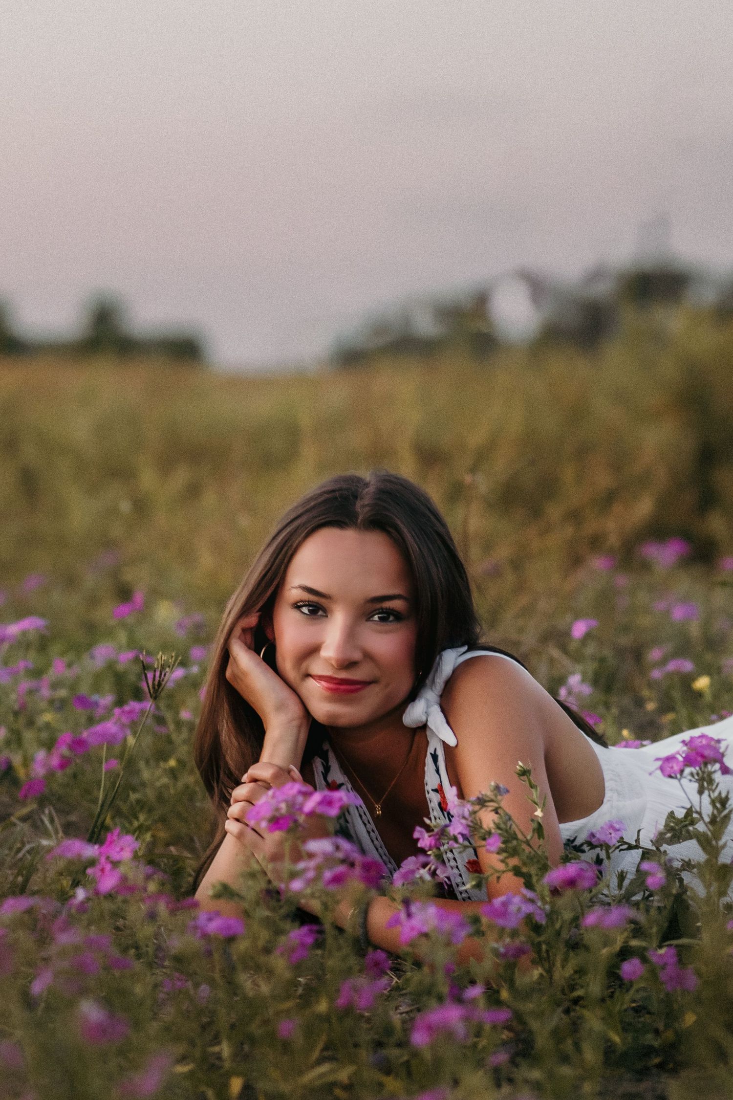 A person poses while lying in a field of purple flowers at dusk.
