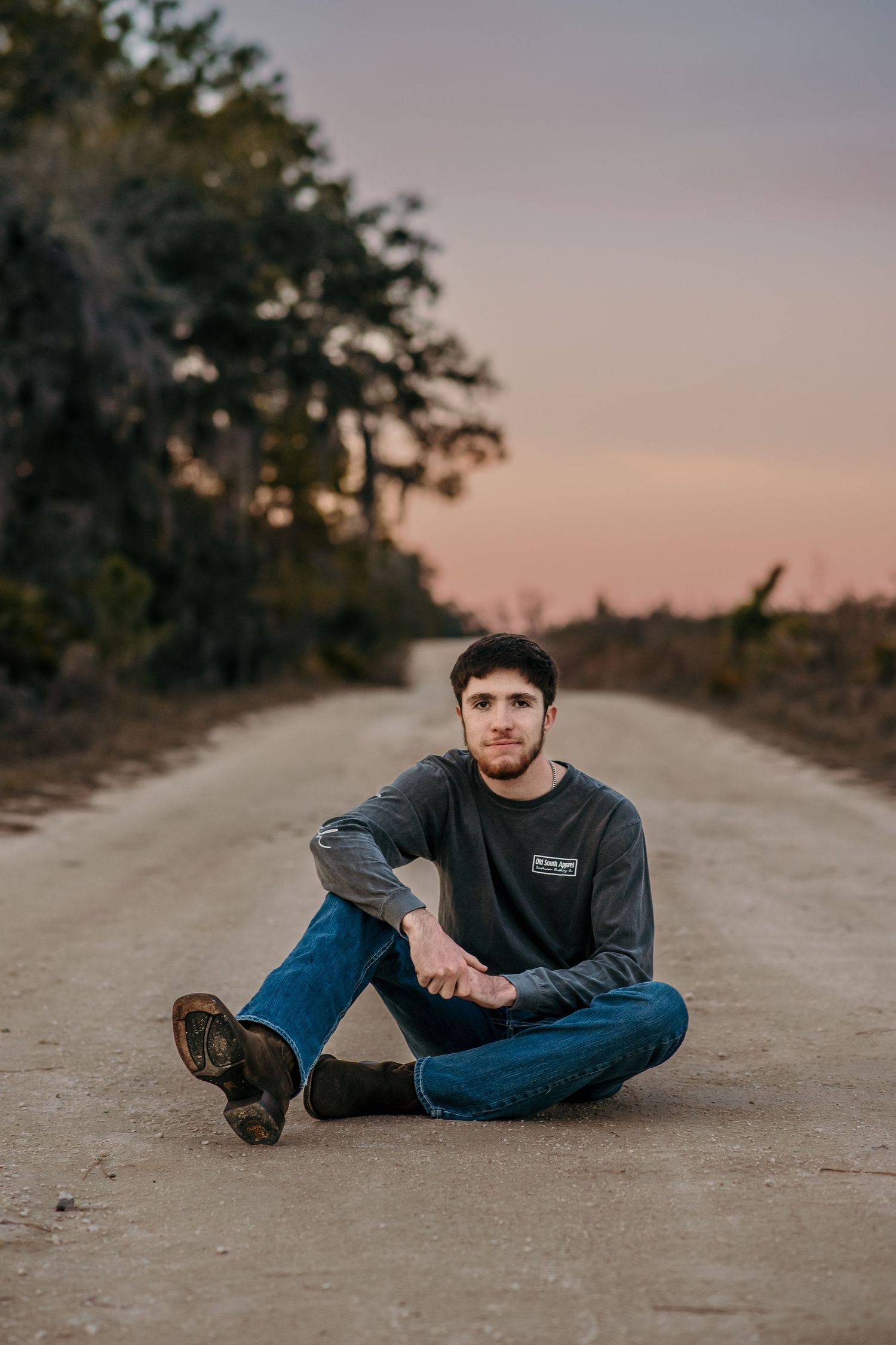A casual outdoor portrait of someone sitting on a dirt road at dusk wearing jeans and a sweatshirt.