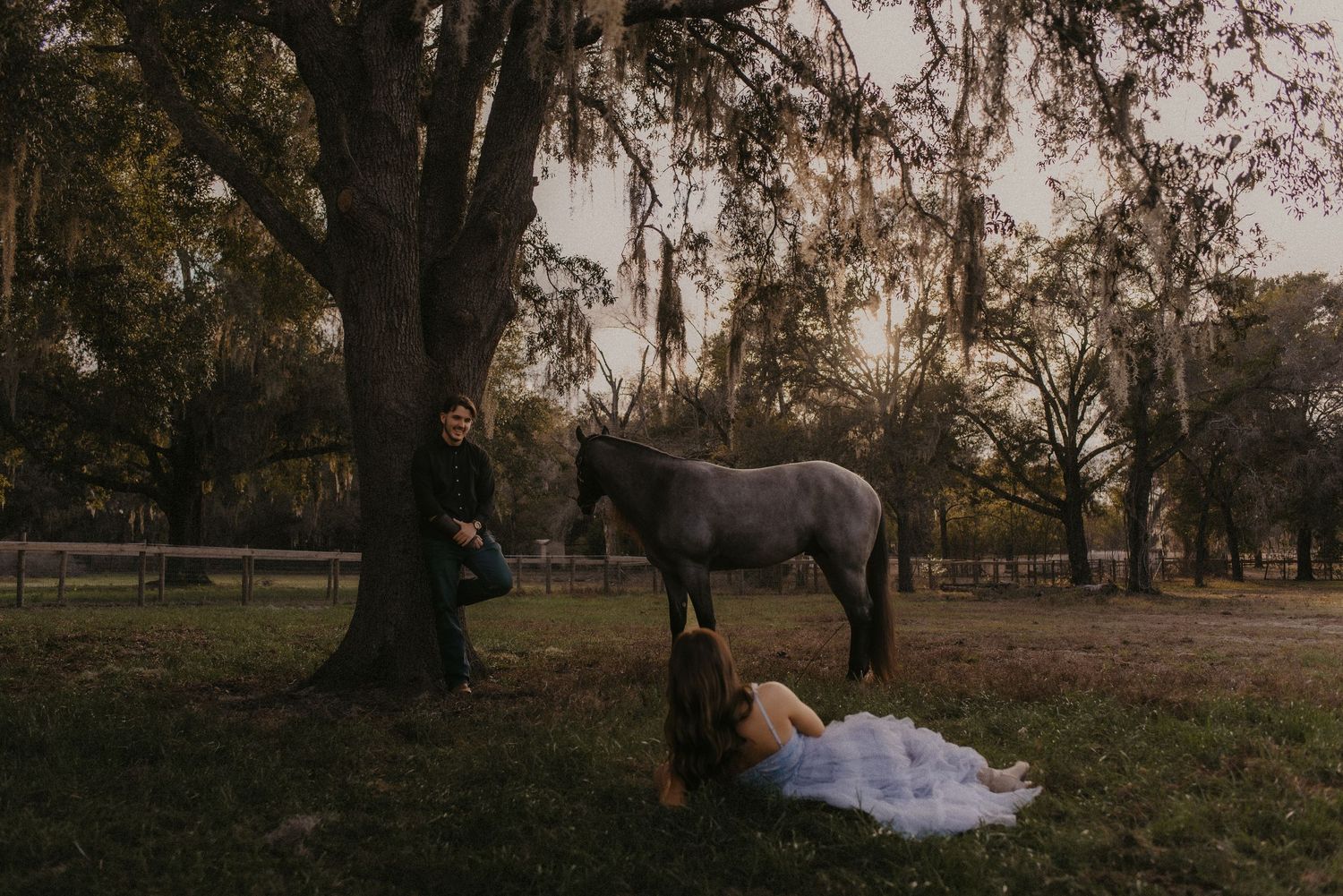 A couple in formal wedding attire poses romantically in a rustic outdoor setting with autumn trees at sunset.