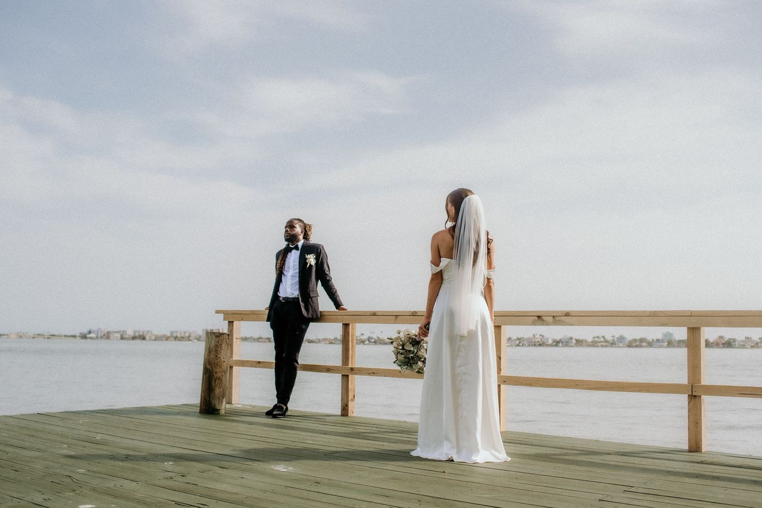 A couple in wedding attire stands together on a wooden boardwalk overlooking a serene waterfront landscape.