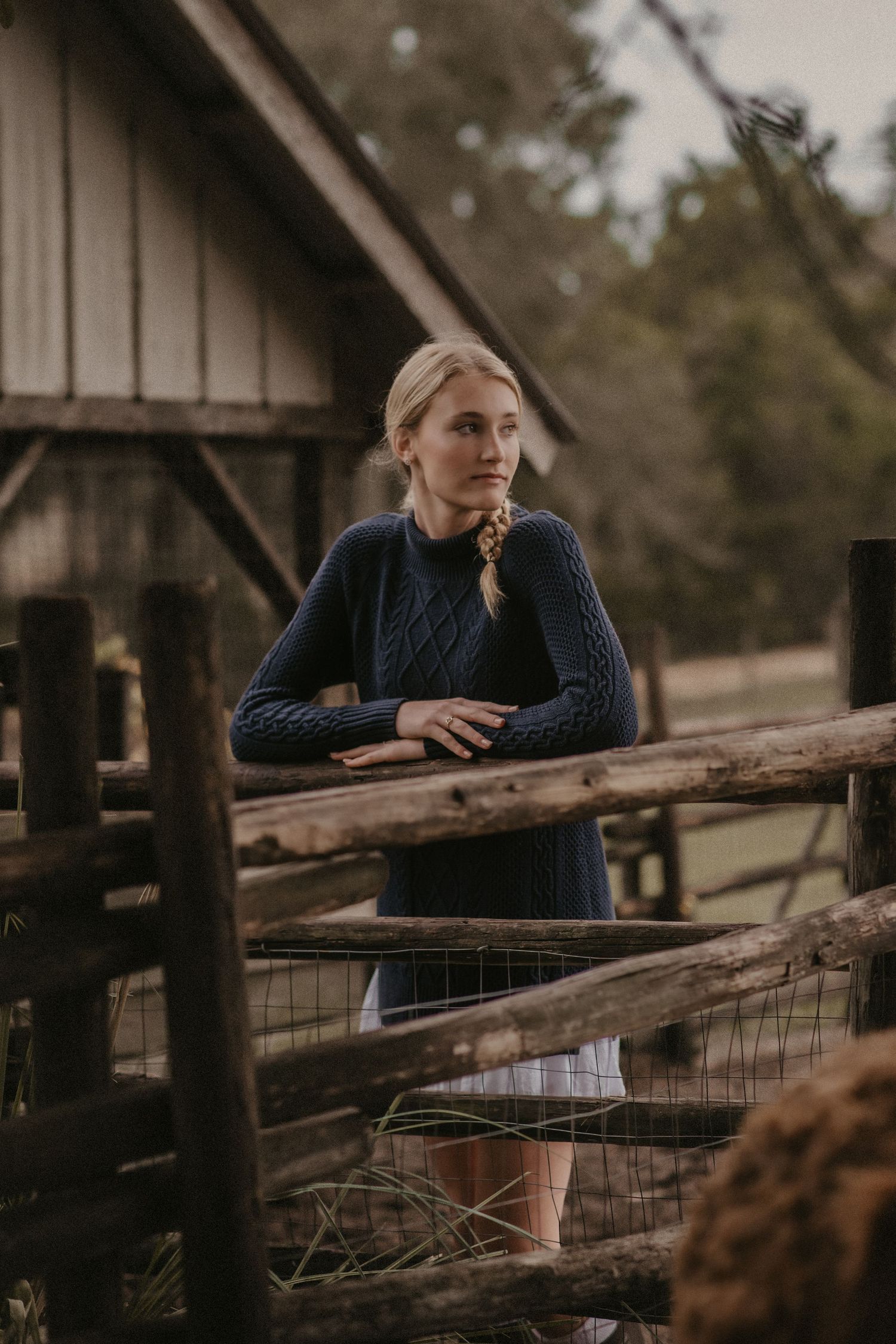 Person in navy sweater leans against rustic wooden fence at dusk with barn in background.