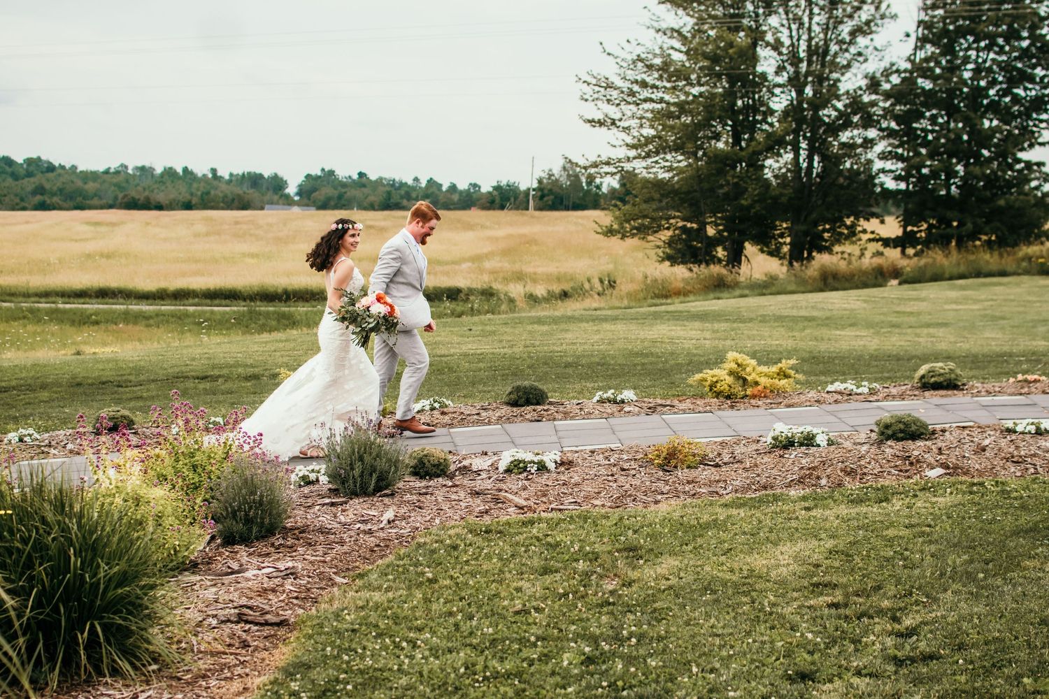 A couple walks along a landscaped garden path in wedding attire.