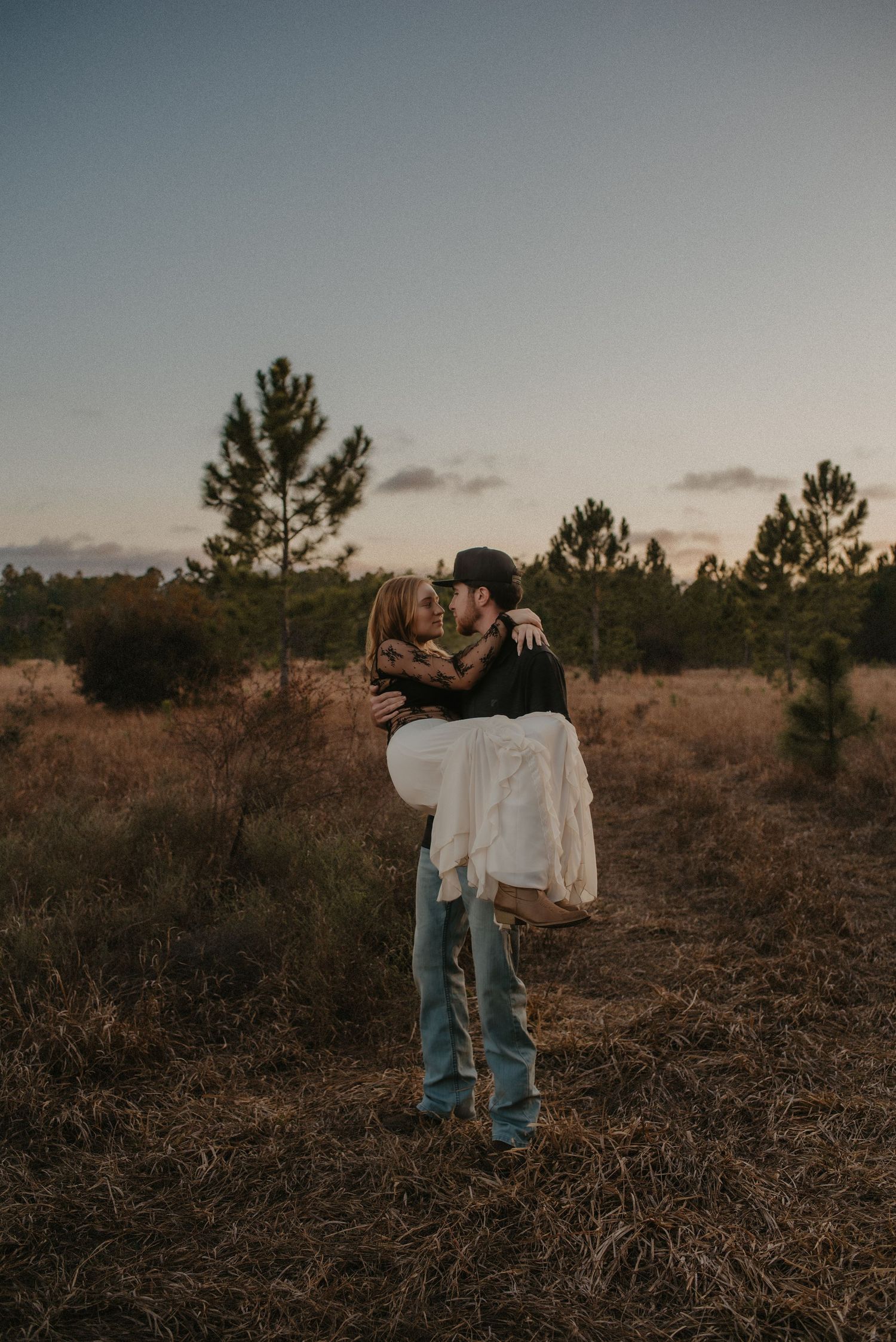 A couple in casual attire embraces during sunset in a rustic field with scattered pine trees in the background.