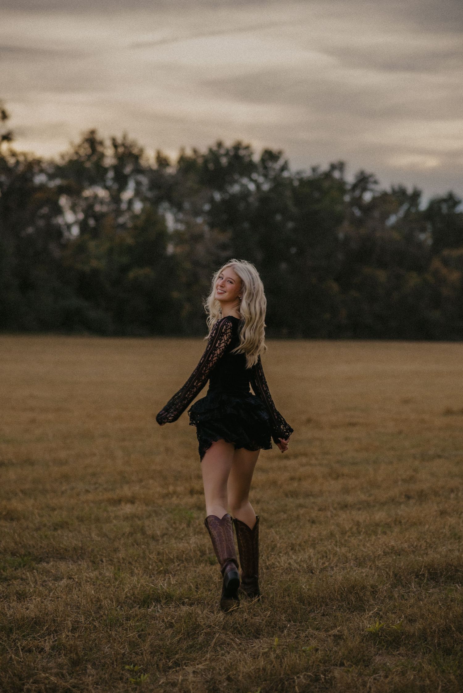 A person in black clothing dances freely in a golden field at sunset under moody clouds.