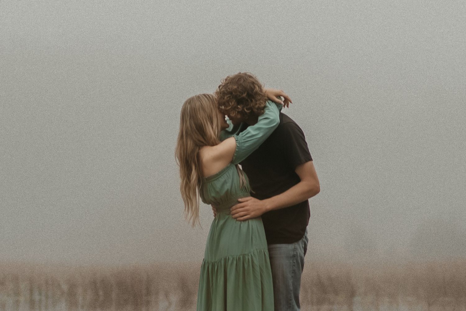 A couple in black and sage green outfits embrace in a romantic outdoor photoshoot with a misty background.