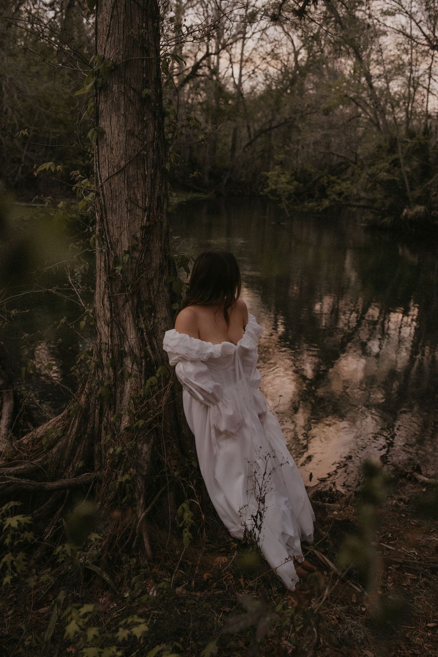 Ethereal figure in flowing white dress stands among cypress trees at twilight beside a misty swamp.