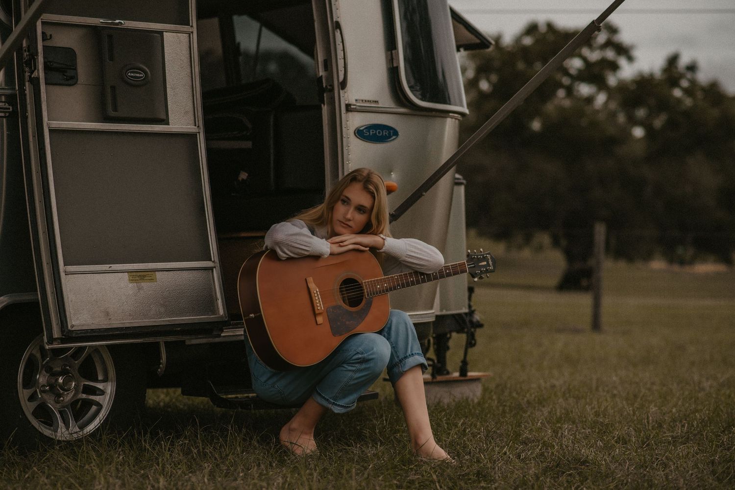 Someone sits barefoot outside an RV playing an acoustic guitar in a grassy field at dusk.