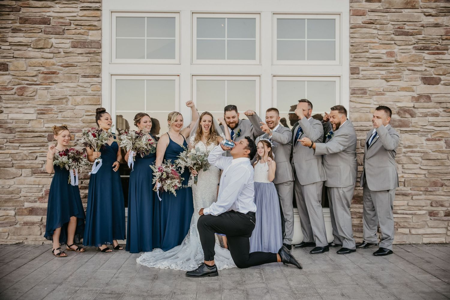 Wedding party poses playfully outside a stone building wearing navy and gray attire.