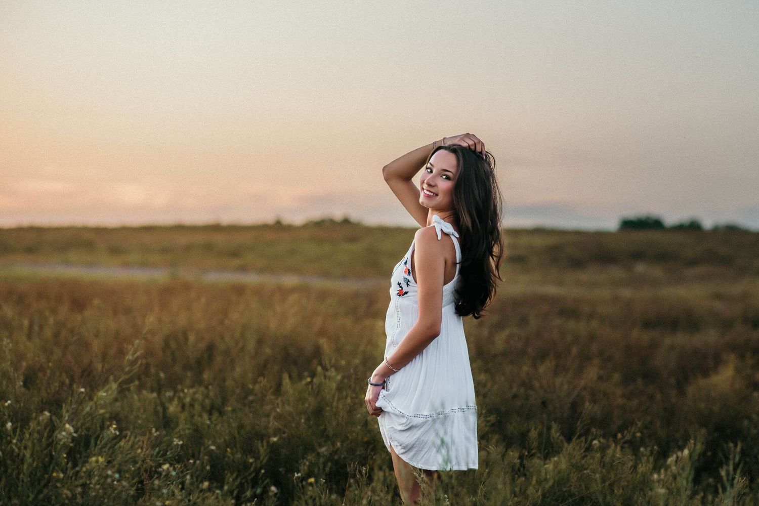 Figure in white dress stands gracefully against sunset sky in meadow.