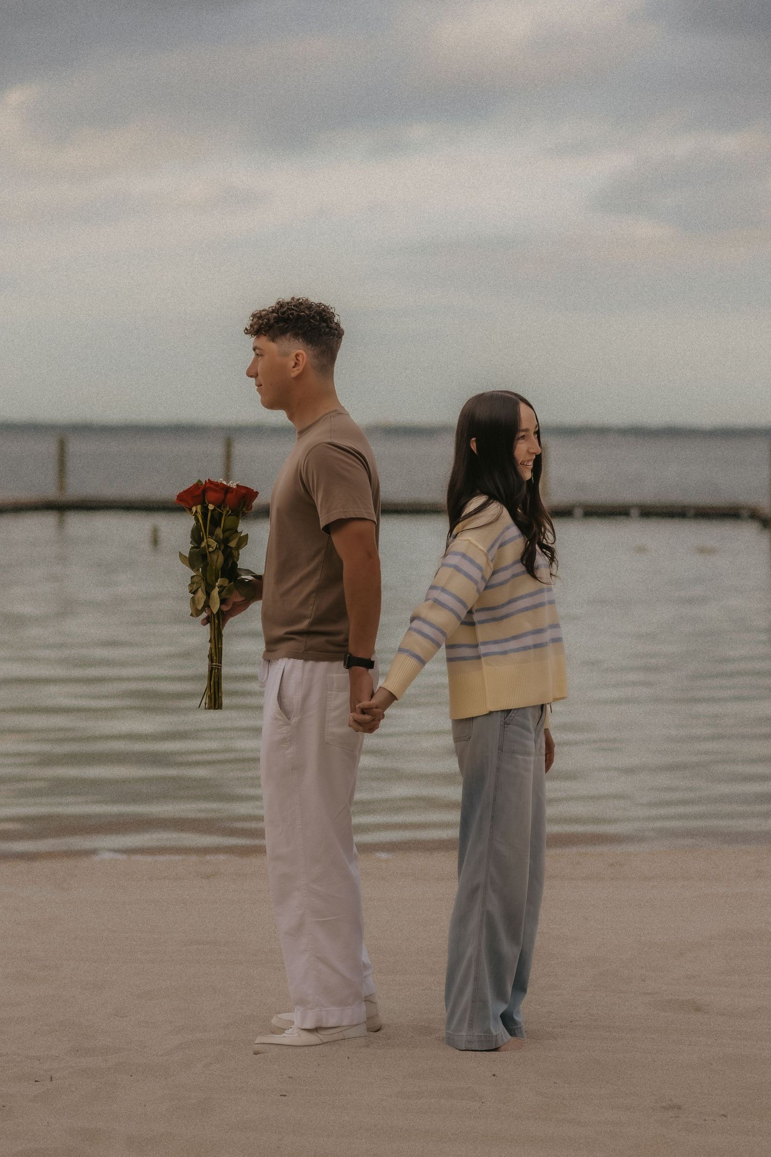 A couple stands back-to-back on a beach at sunset, with one person holding red roses while holding hands.