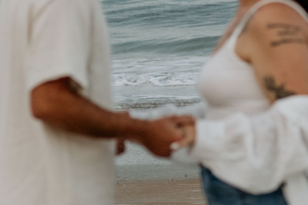 Close-up detail of couple holding hands on beach with ocean waves in background.