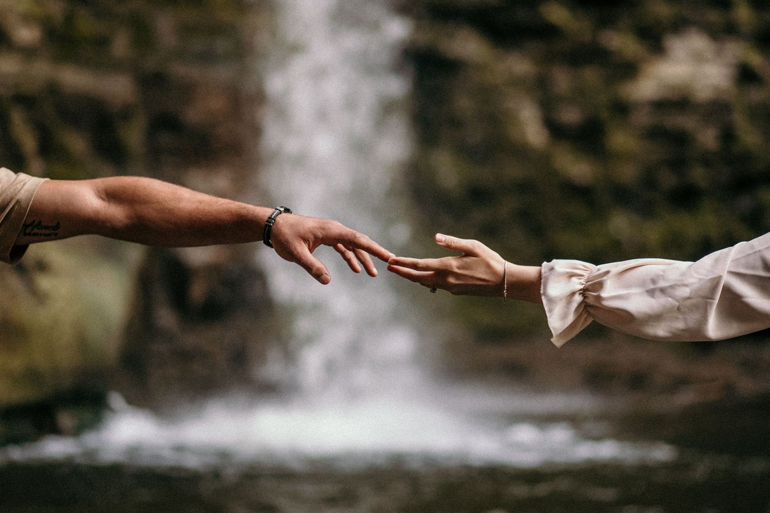 Two hands reaching toward each other in front of a misty waterfall.