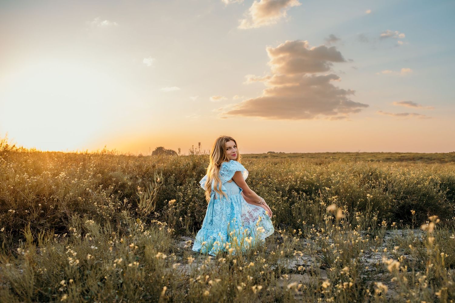 A person in a light blue dress twirls in a golden wildflower field at sunset.