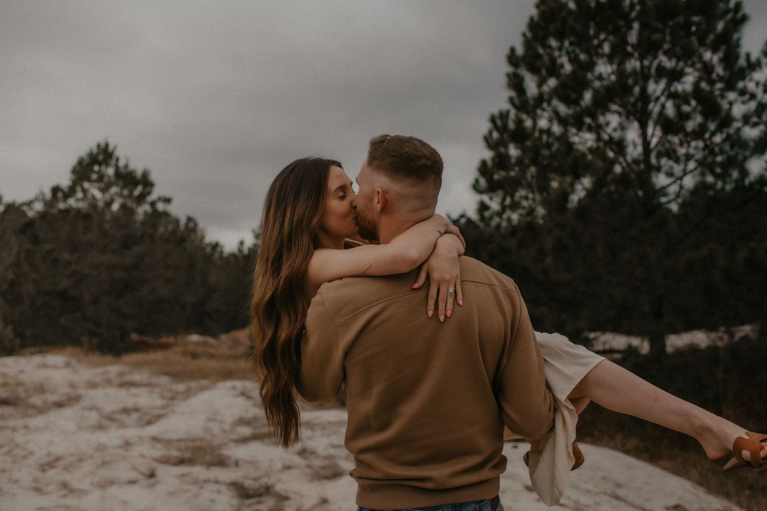 A romantic couple embraces in a moody outdoor setting with pine trees and overcast skies in the background.