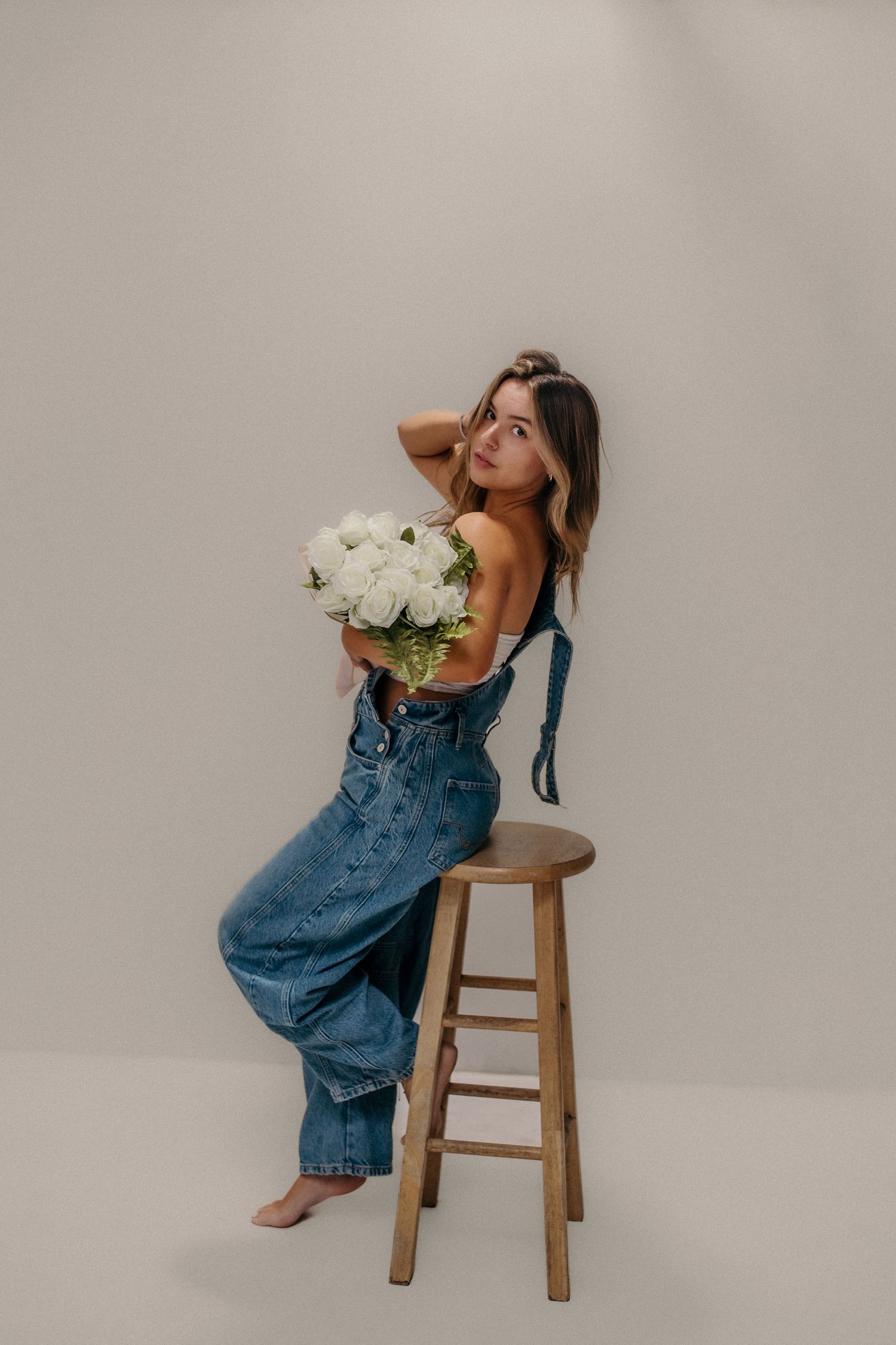 A person in denim overalls poses playfully on a wooden stool against a neutral backdrop with white flowers.