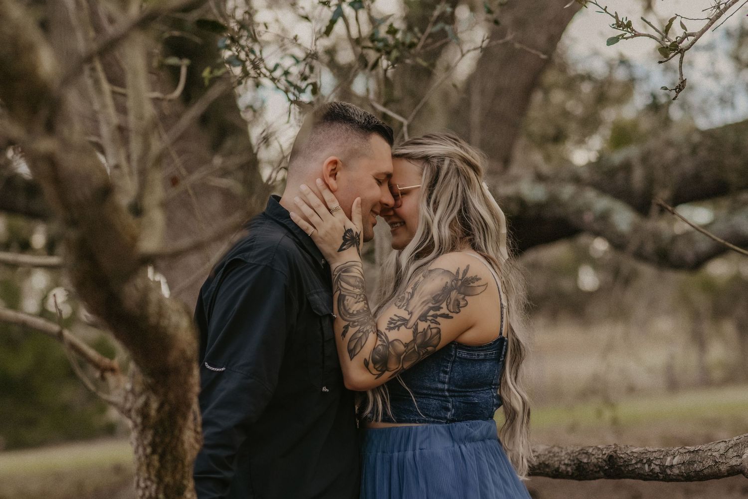 A romantic couple embraces in a natural outdoor setting surrounded by blooming trees and soft sunlight.