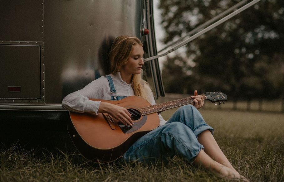 Person playing acoustic guitar while sitting beside vintage vehicle at dusk.
