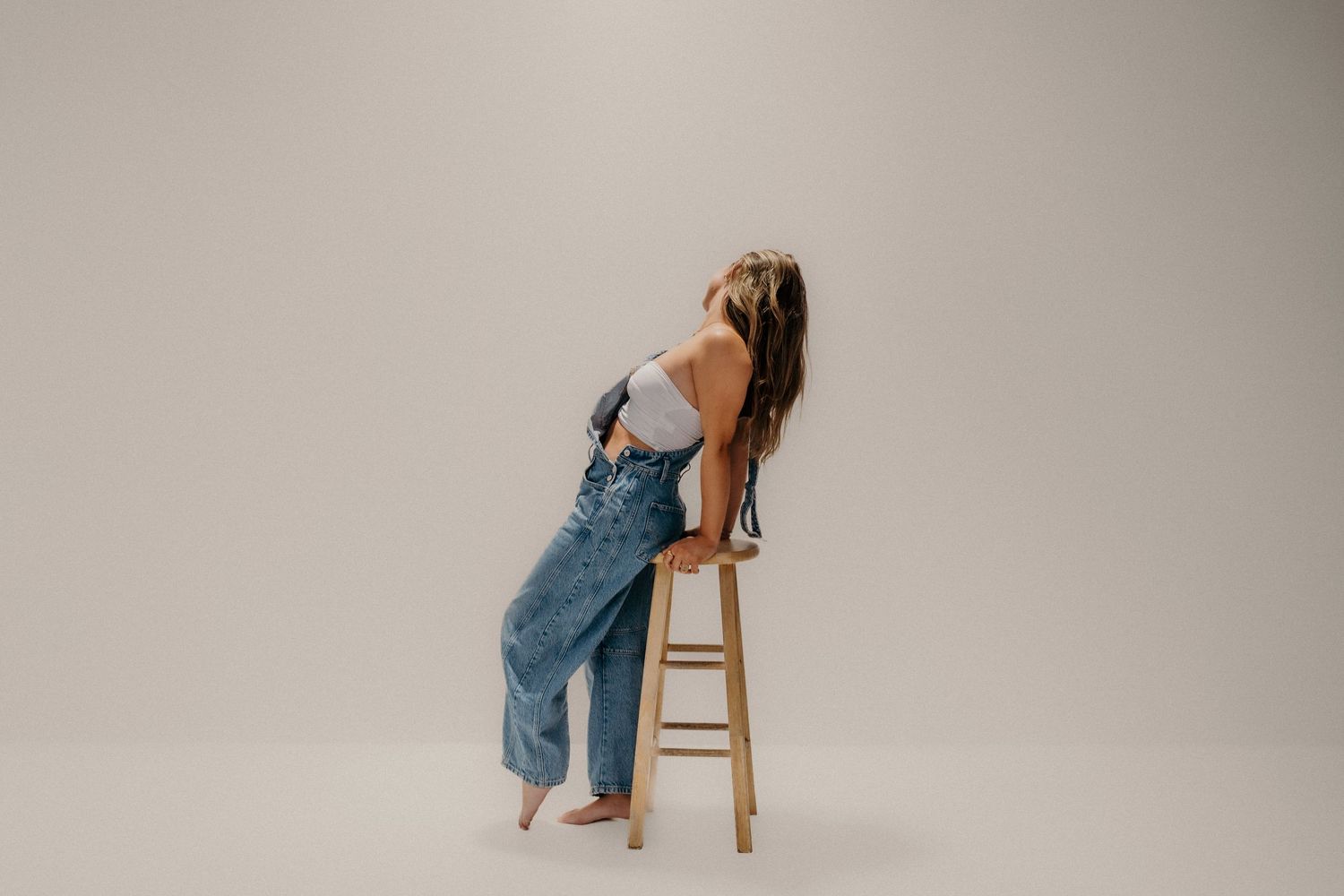 A person in blue denim overalls sits on a wooden stool against a neutral background in various artistic poses.