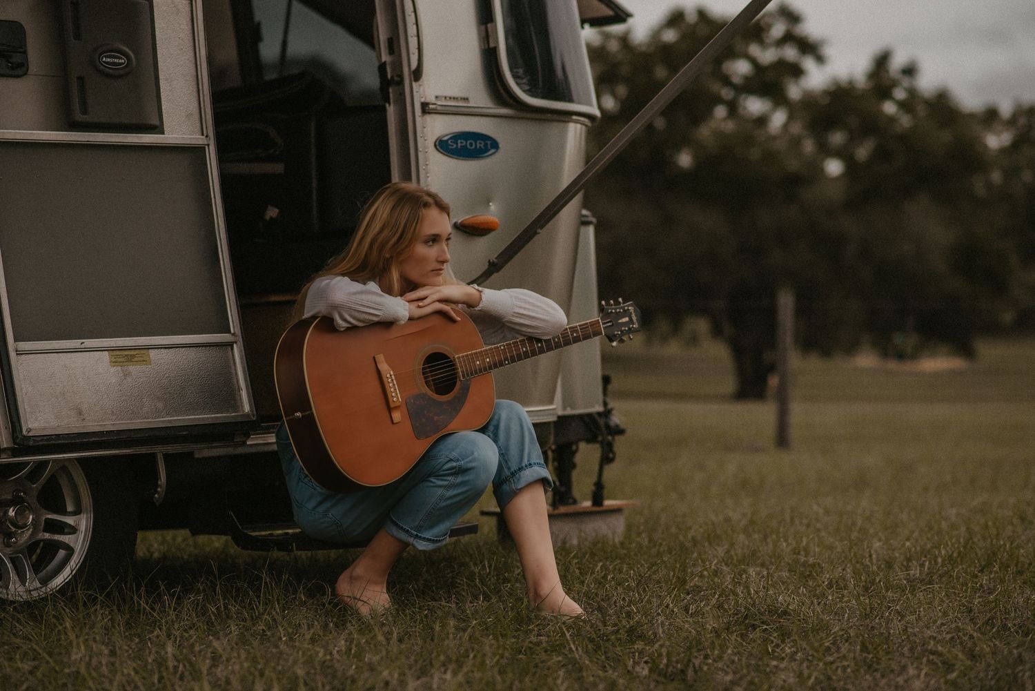 A musician sits barefoot at the entrance of an RV with an acoustic guitar on a grassy field.