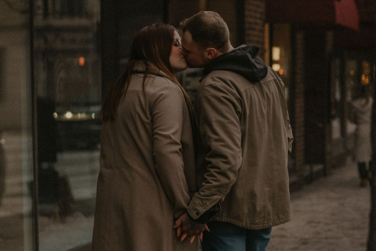 Two people in winter coats walk down a dimly lit urban street with brick buildings and storefronts at night.