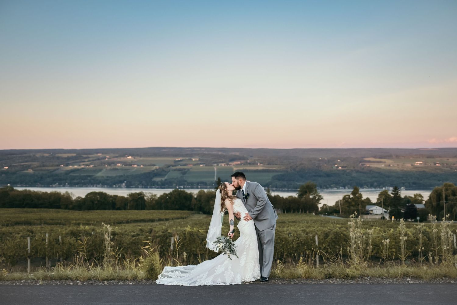 A romantic wedding couple embraces on a hilltop overlooking a scenic lake at sunset.