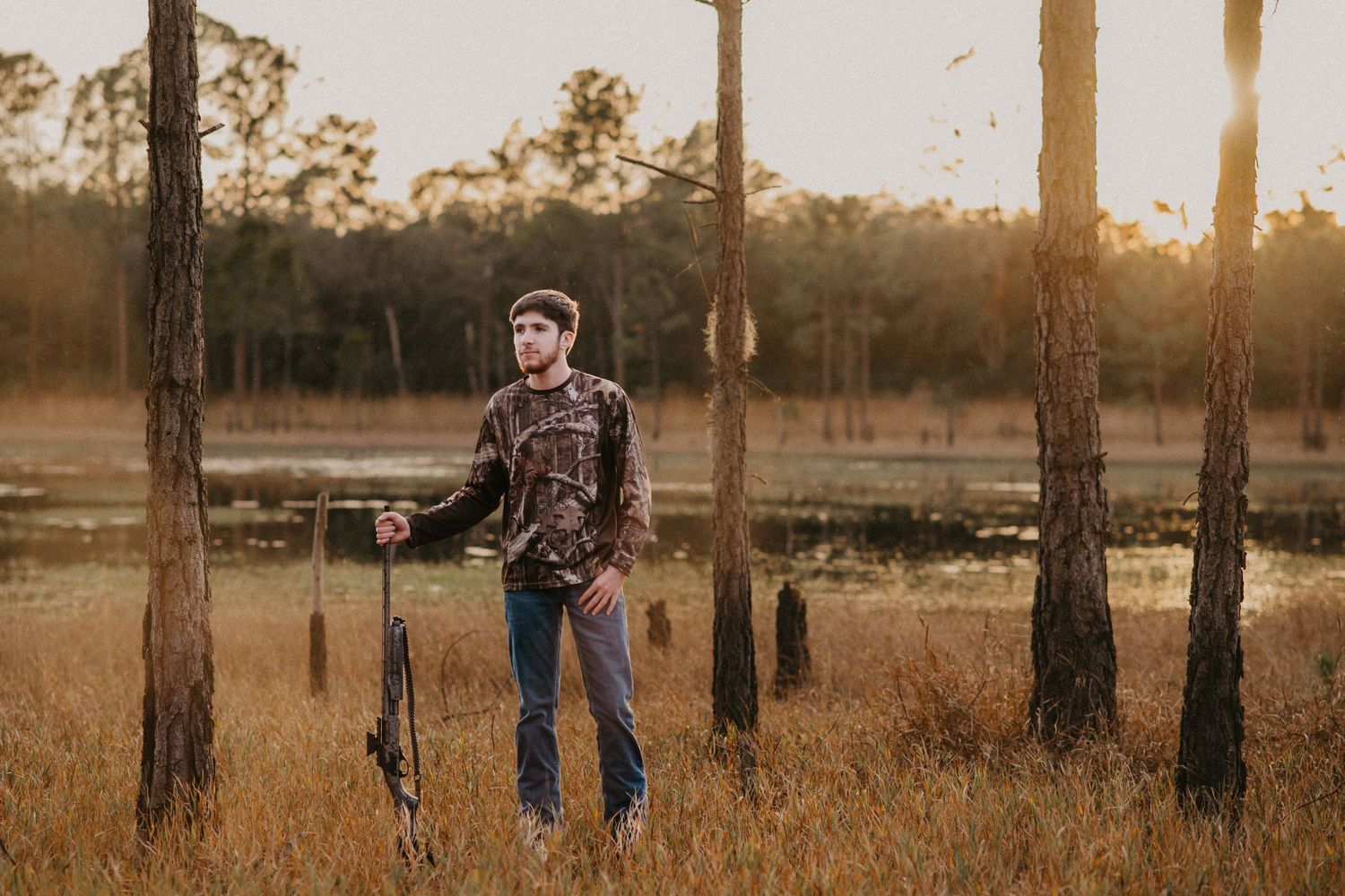 Hunter poses with rifle among tall pine trees during golden hour.