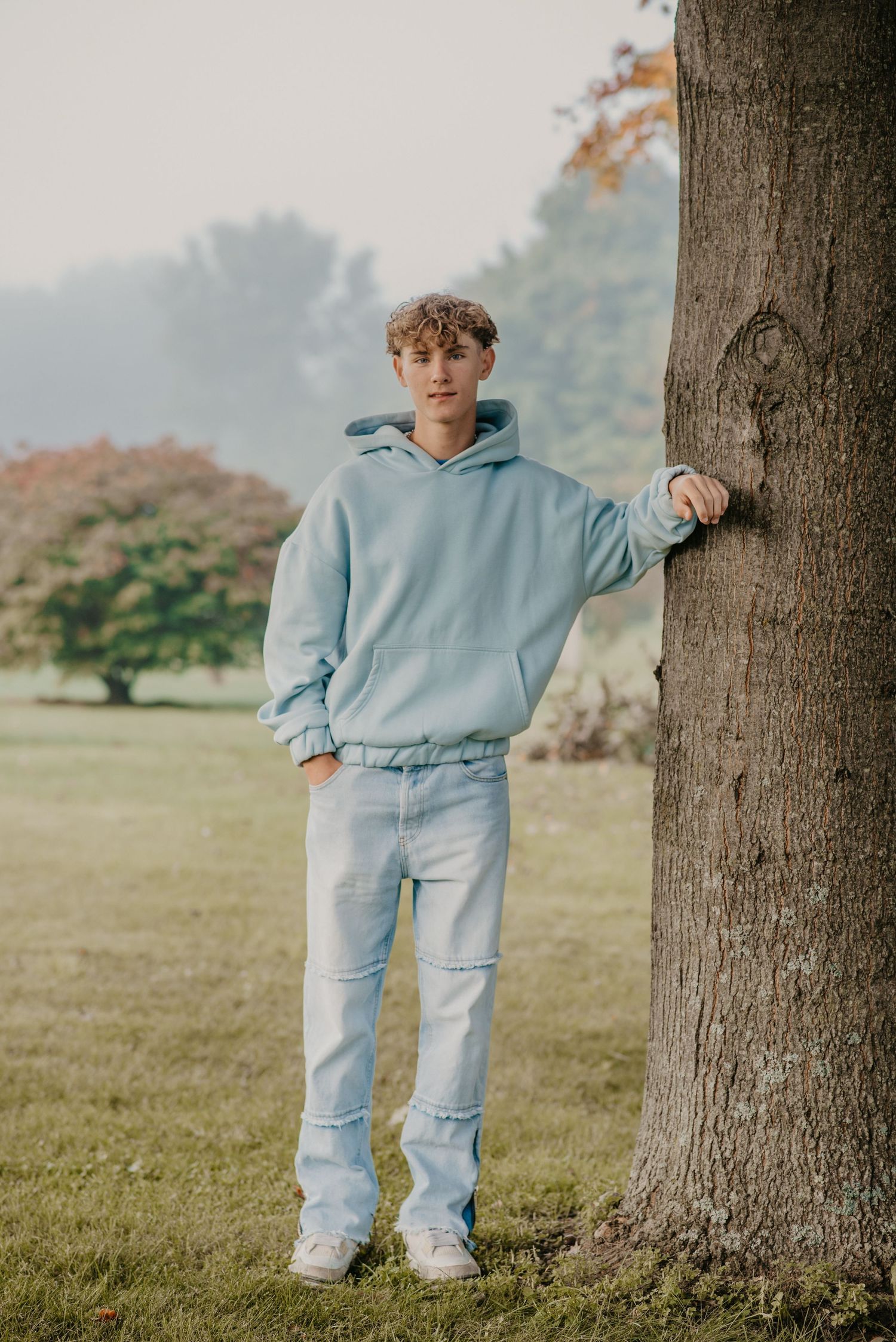 Full length portrait in matching light blue hoodie and jeans against tree in park setting.