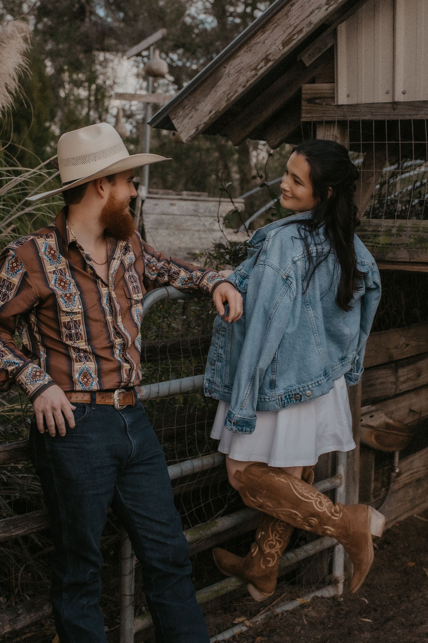 A western-styled couple in denim and cowboy boots pose together near rustic wooden fencing at sunset.