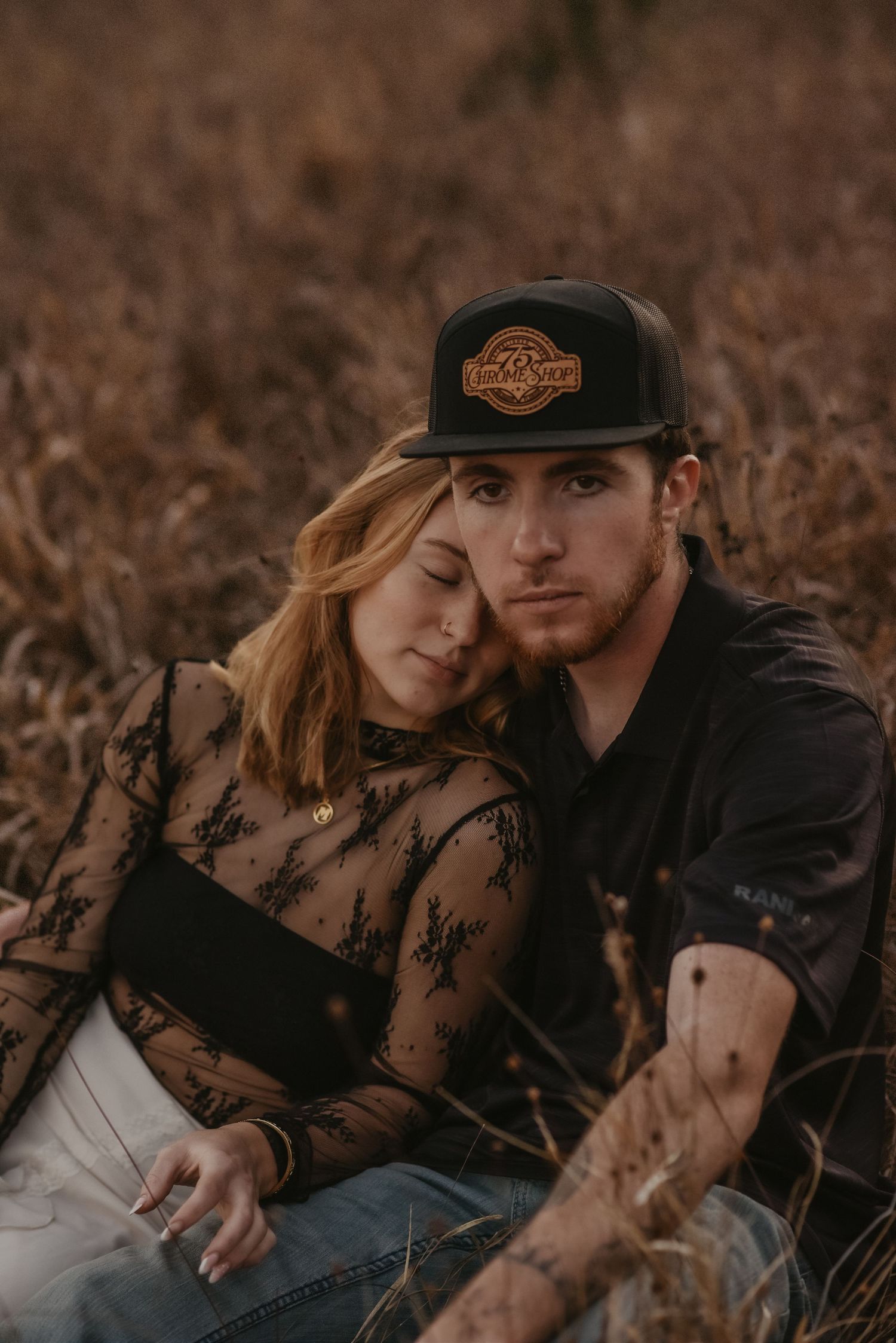 A couple sits intimately in a brown autumn field at sunset, wearing casual clothing and a baseball cap.