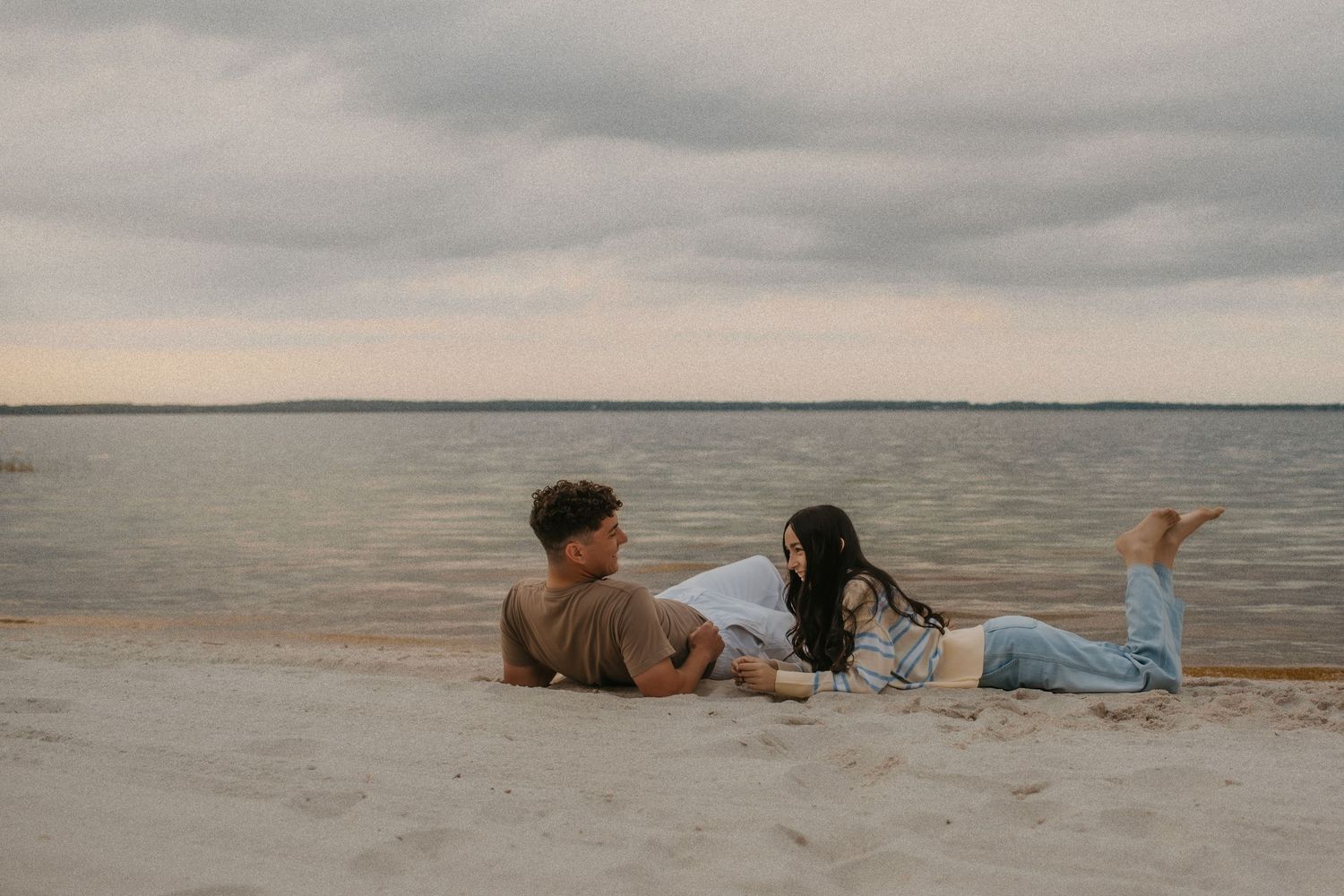 Two people lying on a sandy beach at sunset looking out over calm waters under cloudy skies.