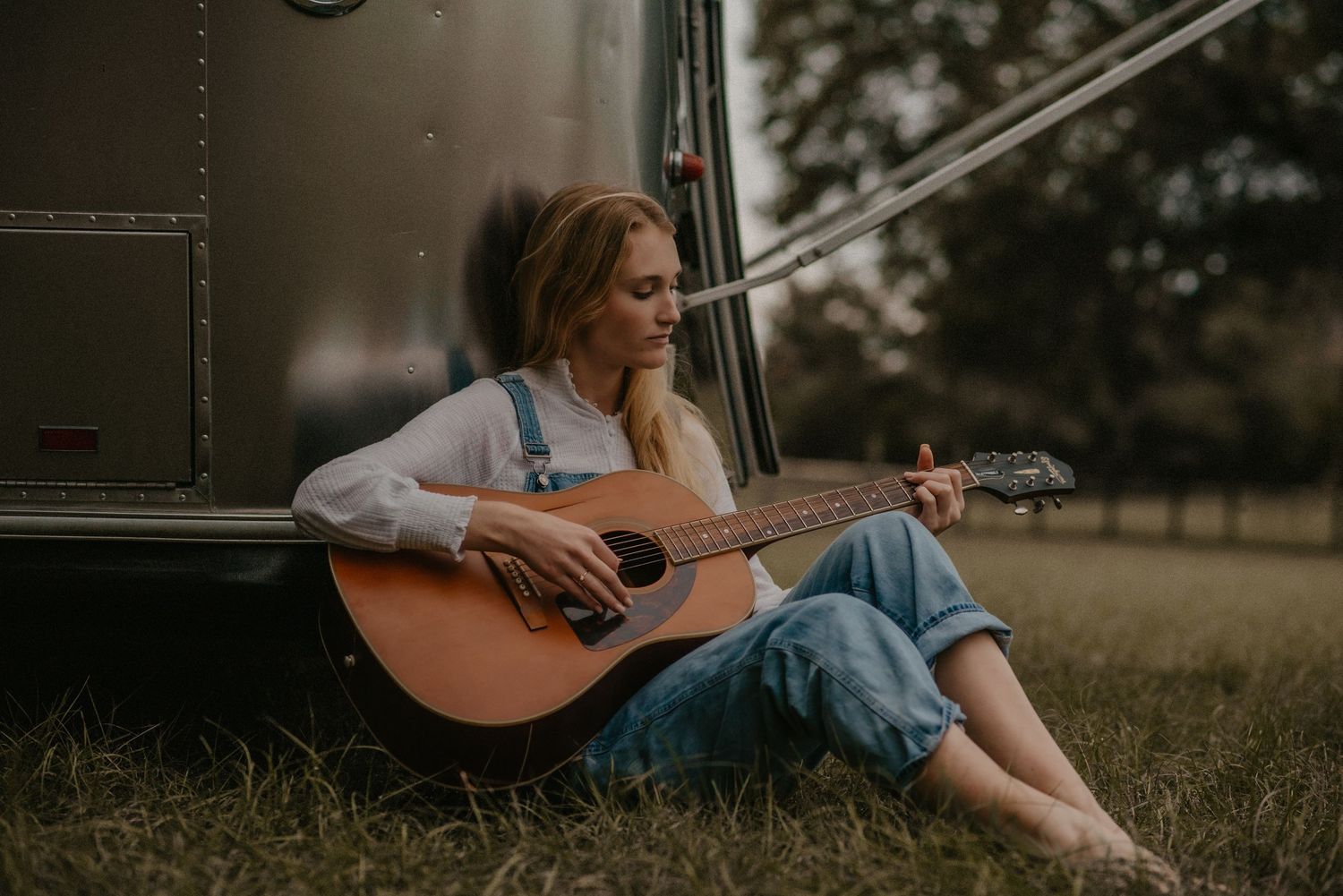 Person in casual denim sits on grass playing acoustic guitar in moody outdoor setting at sunset.