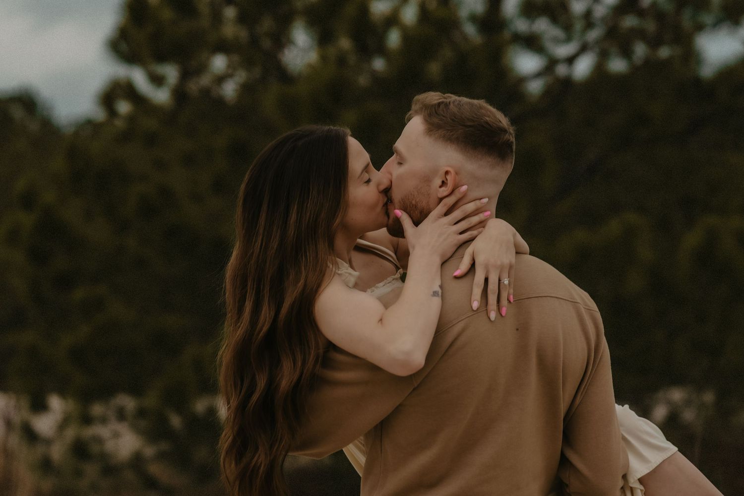 A romantic couple embraces and kisses outdoors during golden hour with trees in the background.
