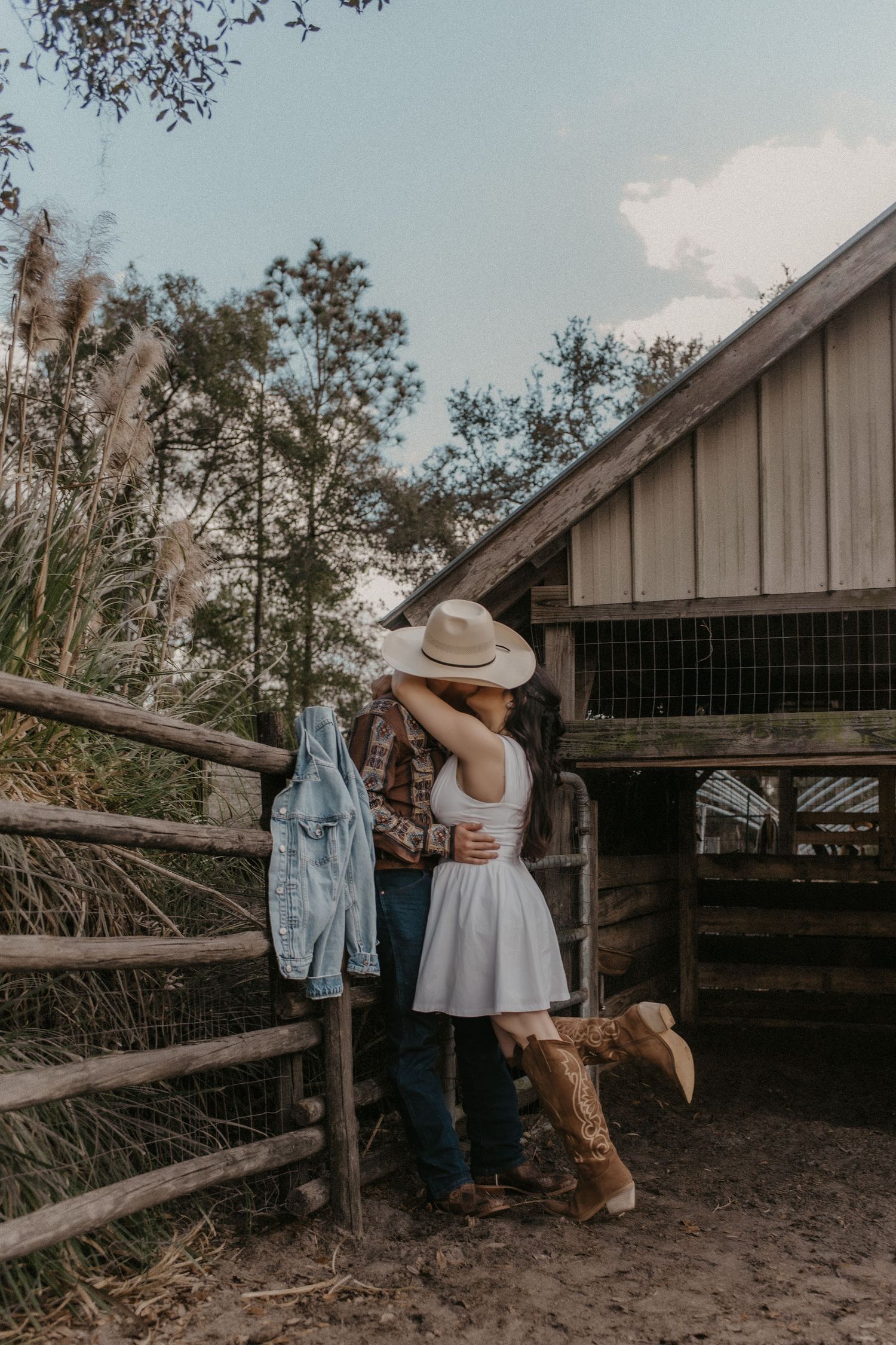 A romantic couple in western attire leans against a rustic wooden fence at sunset near a barn.