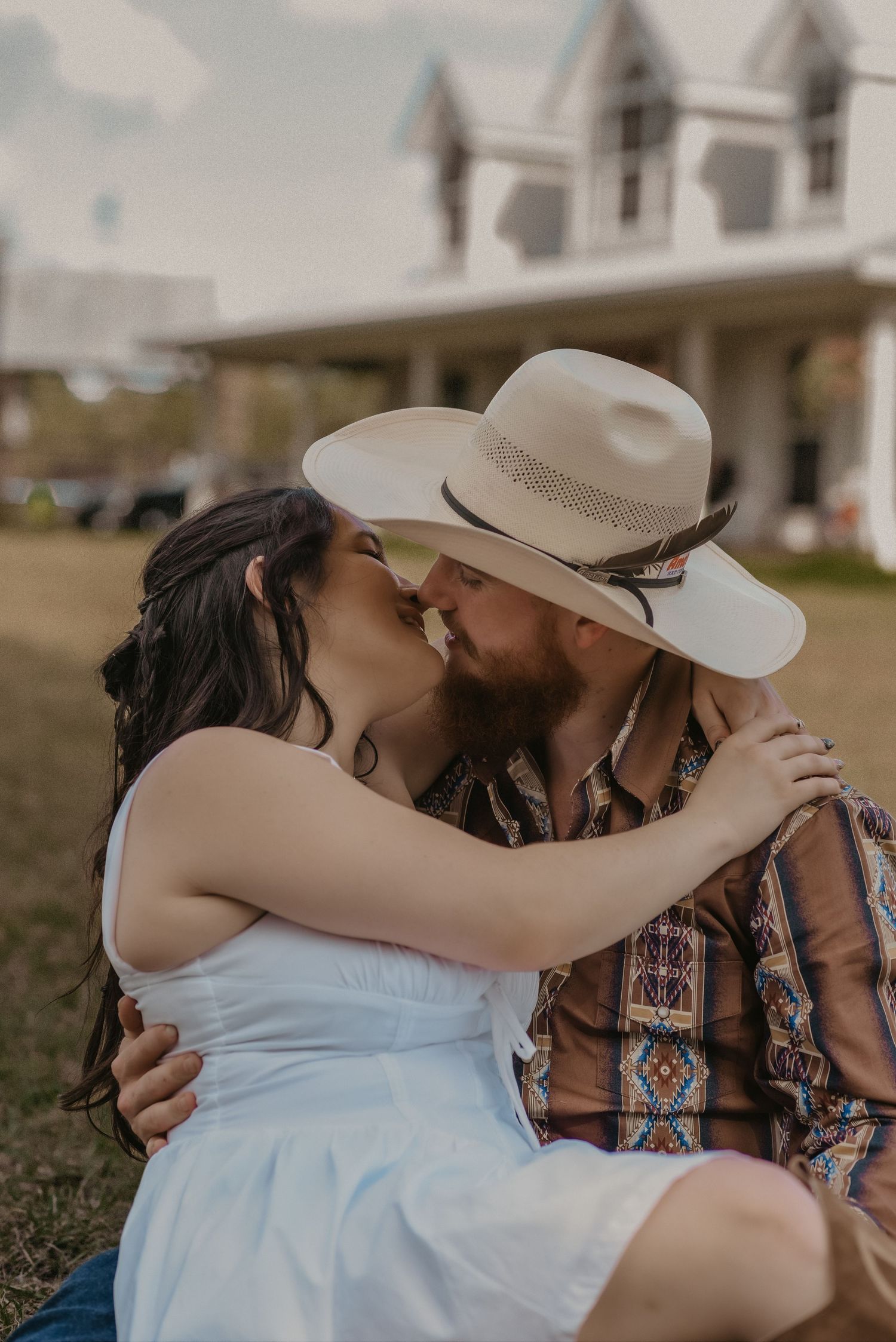 A romantic couple embraces and kisses outdoors with a rustic farmhouse in the background at sunset.