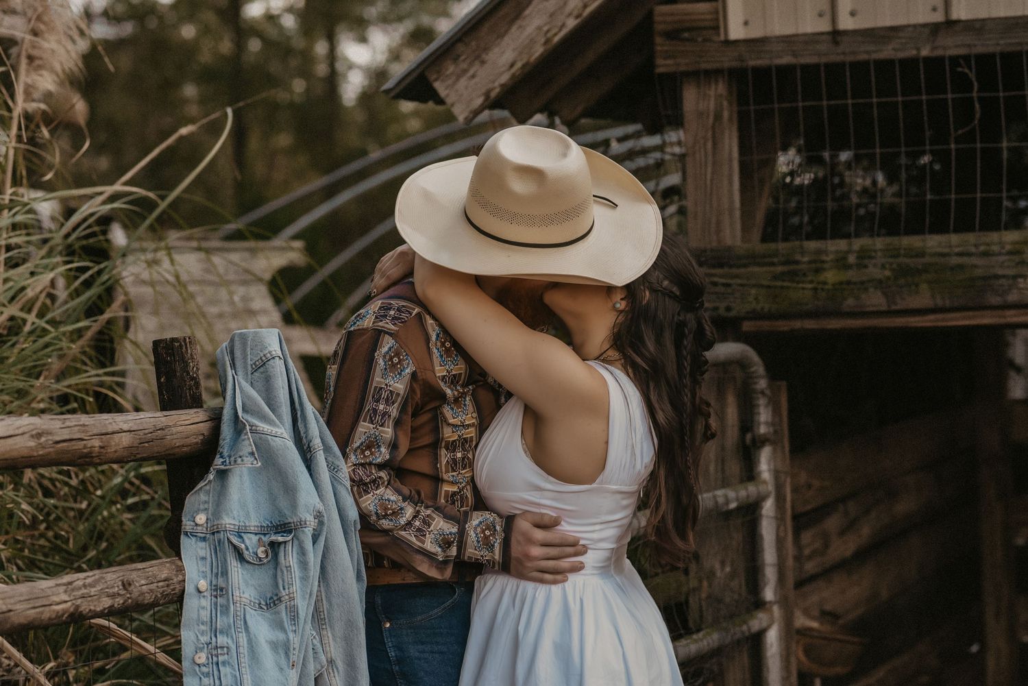 A romantic country scene with a cowboy hat, plaid shirt and white dress against a rustic wooden fence at sunset.