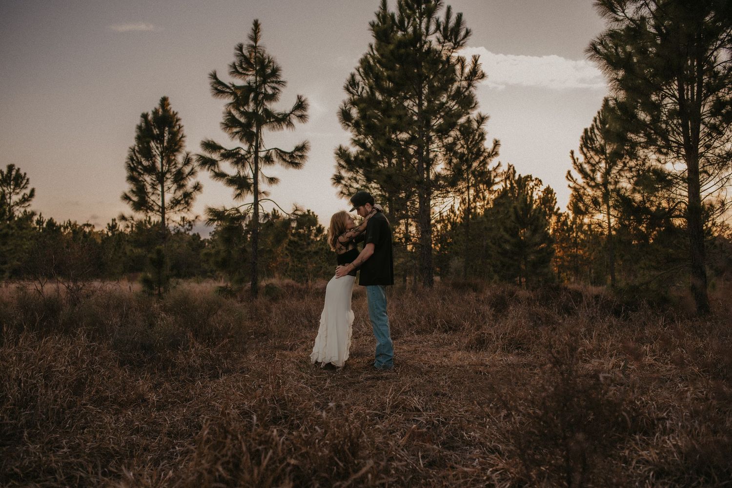 A couple stands together in a field at sunset with tall pine trees silhouetted against the golden sky.