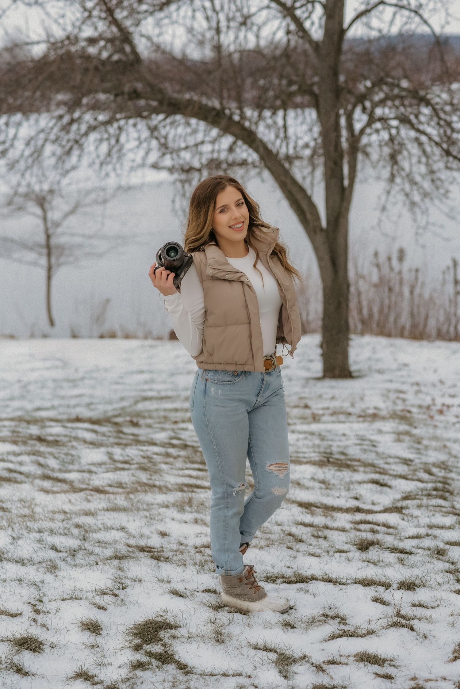 A person in a beige jacket and jeans walks through a snowy winter landscape with bare trees in the background.