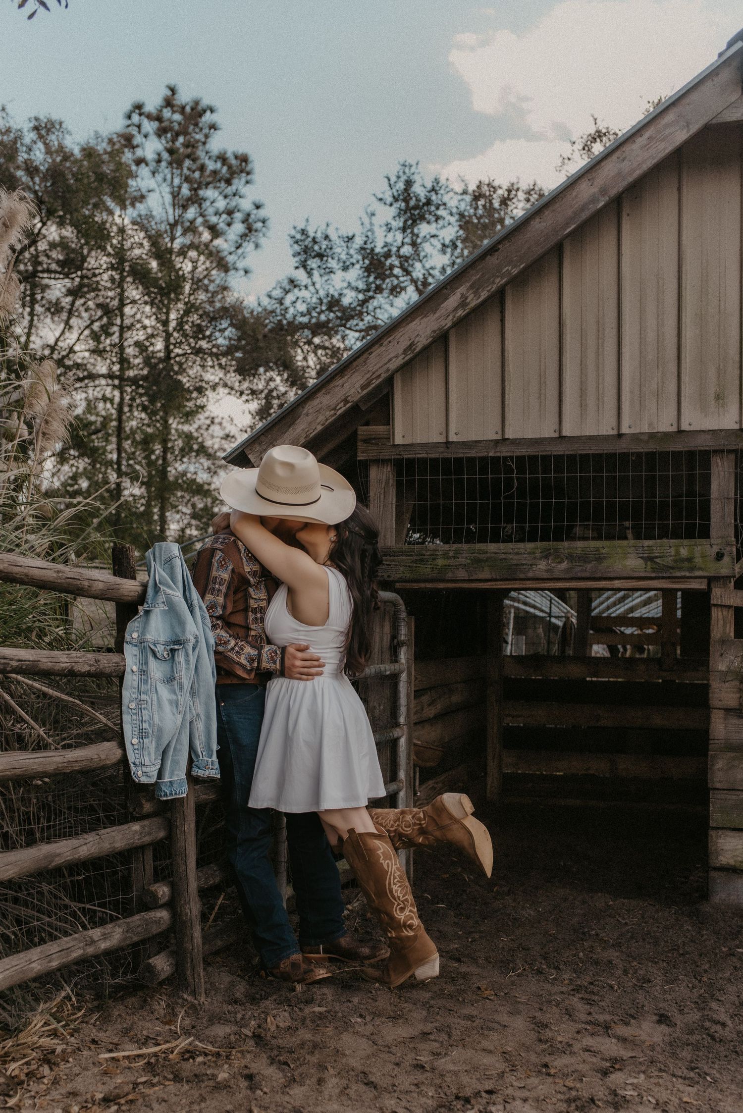 Couple in rustic setting leaning against wooden barn fence during sunset photoshoot.