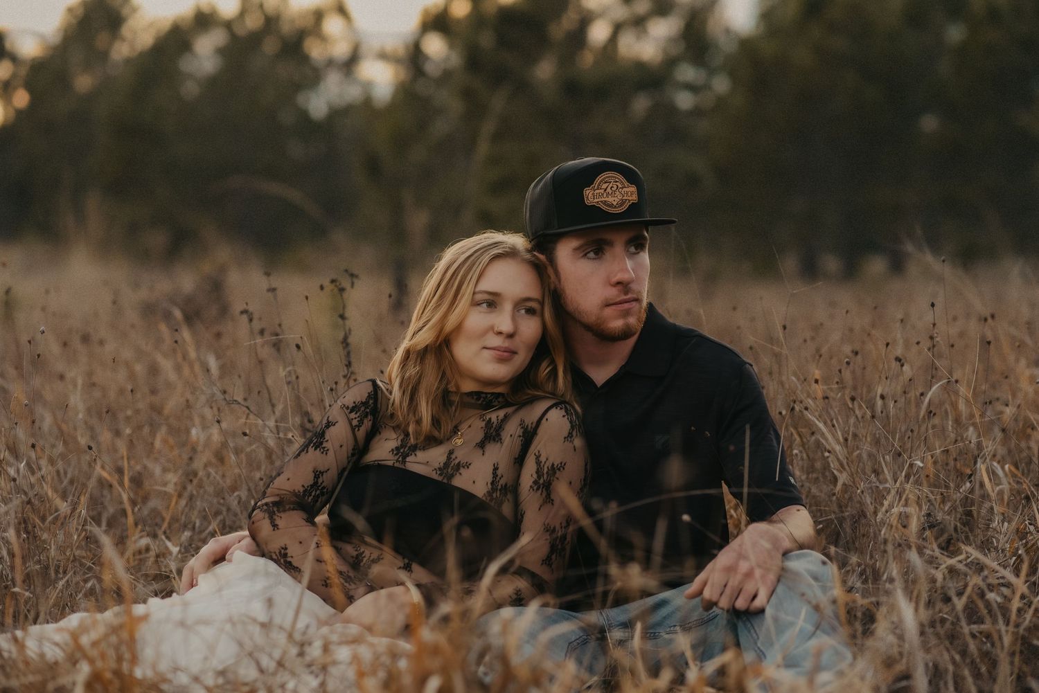 A couple sits together in a golden autumn field during sunset, creating a romantic outdoor scene.