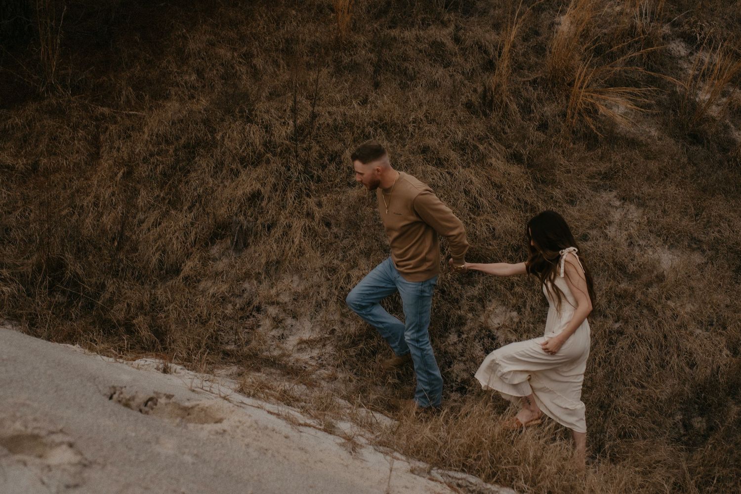 Two people climb up a steep dirt path together, with one wearing a flowing white dress and the other in jeans.