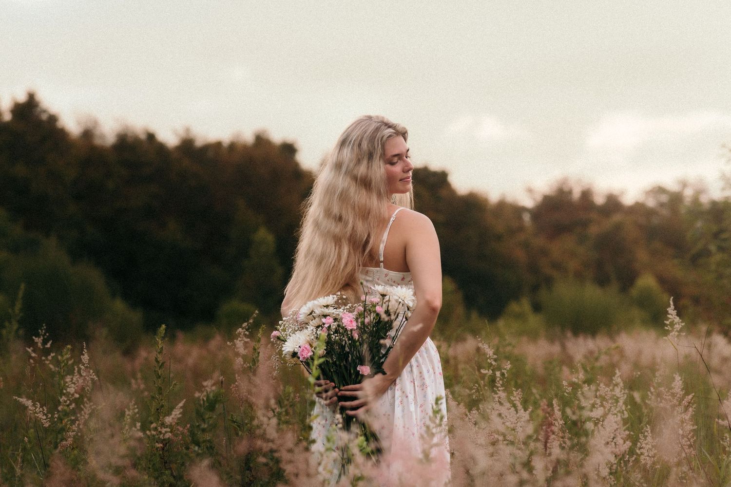 A bride in a white lace wedding dress stands in a golden field at sunset holding a bouquet of flowers.